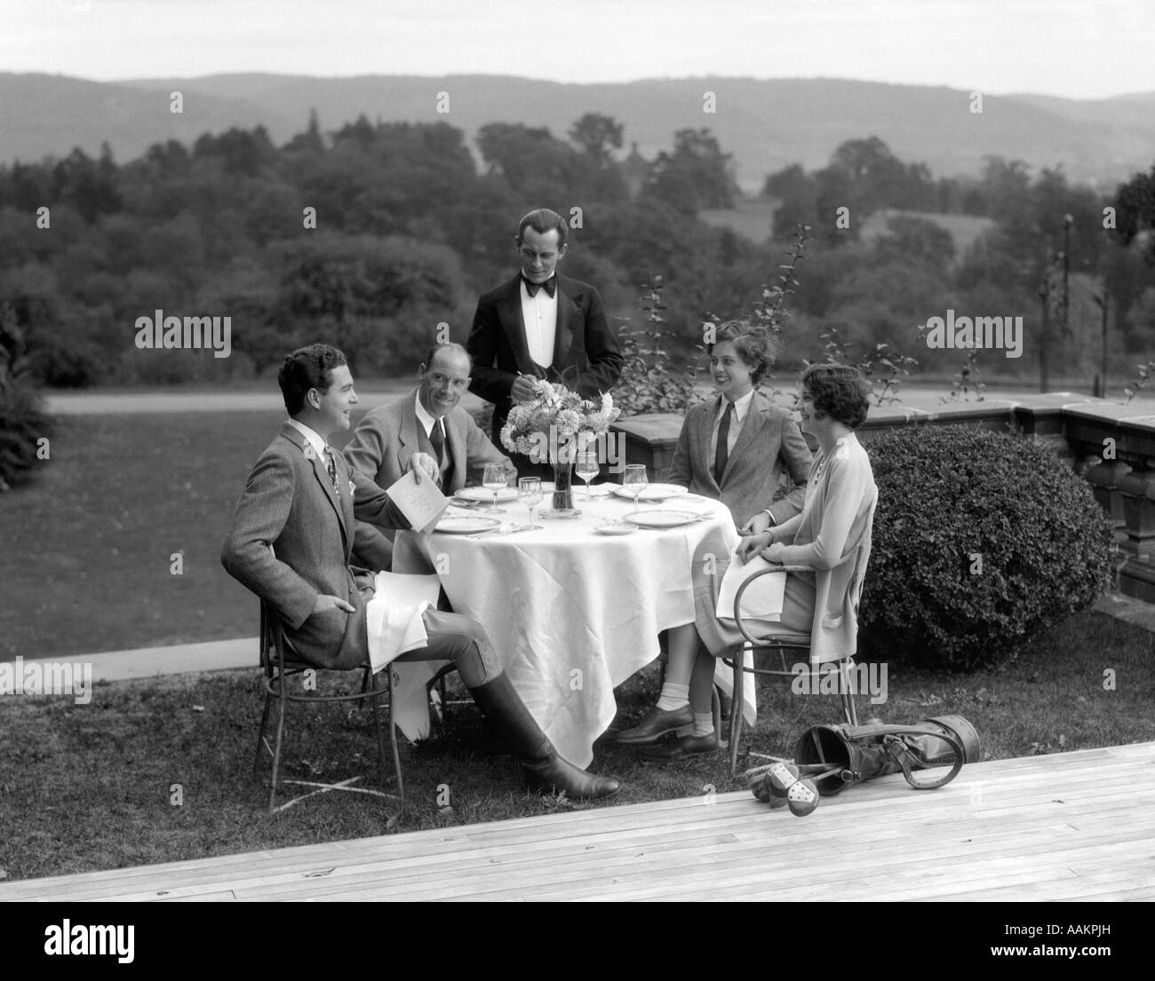 1920s 1930s COUNTRY CLUB SCENE WITH TWO COUPLES WITH GOLF CLUBS HAVING LUNCH OUTDOORS Stock Photo