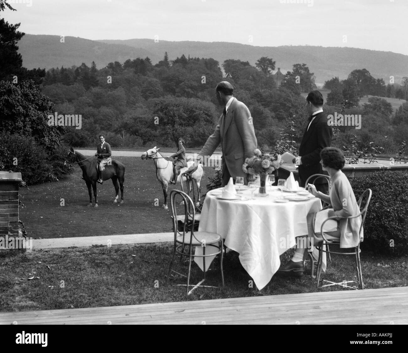 1930s COUNTRY CLUB WITH COUPLE EATING OUTSIDE & ANOTHER COUPLE ON HORSEBACK Stock Photo