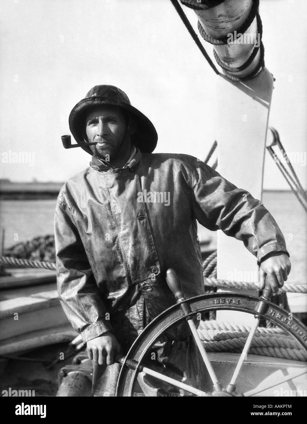 1920s 1930s MAN OLD SALT SAILOR FISHERMAN AT THE HELM OF FISHING BOAT WEARING FOUL WEATHER GEAR AND SMOKING A PIPE Stock Photo
