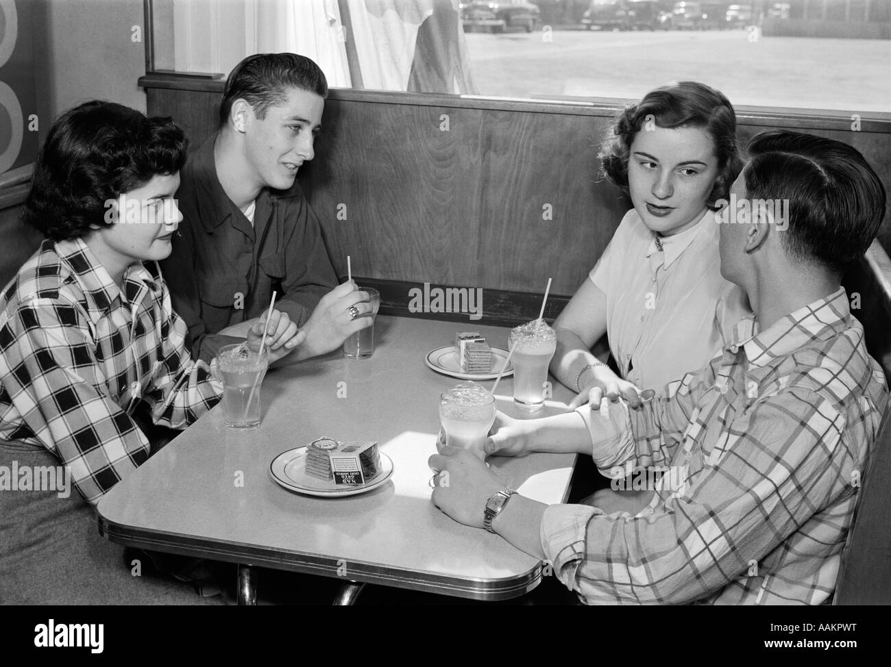 1950s TWO TEENAGE COUPLES AT BOOTH IN DINER WEARING PLAID AND SOLID COLOR SHIRTS DRINKING SODAS TALKING TOGETHER Stock Photo