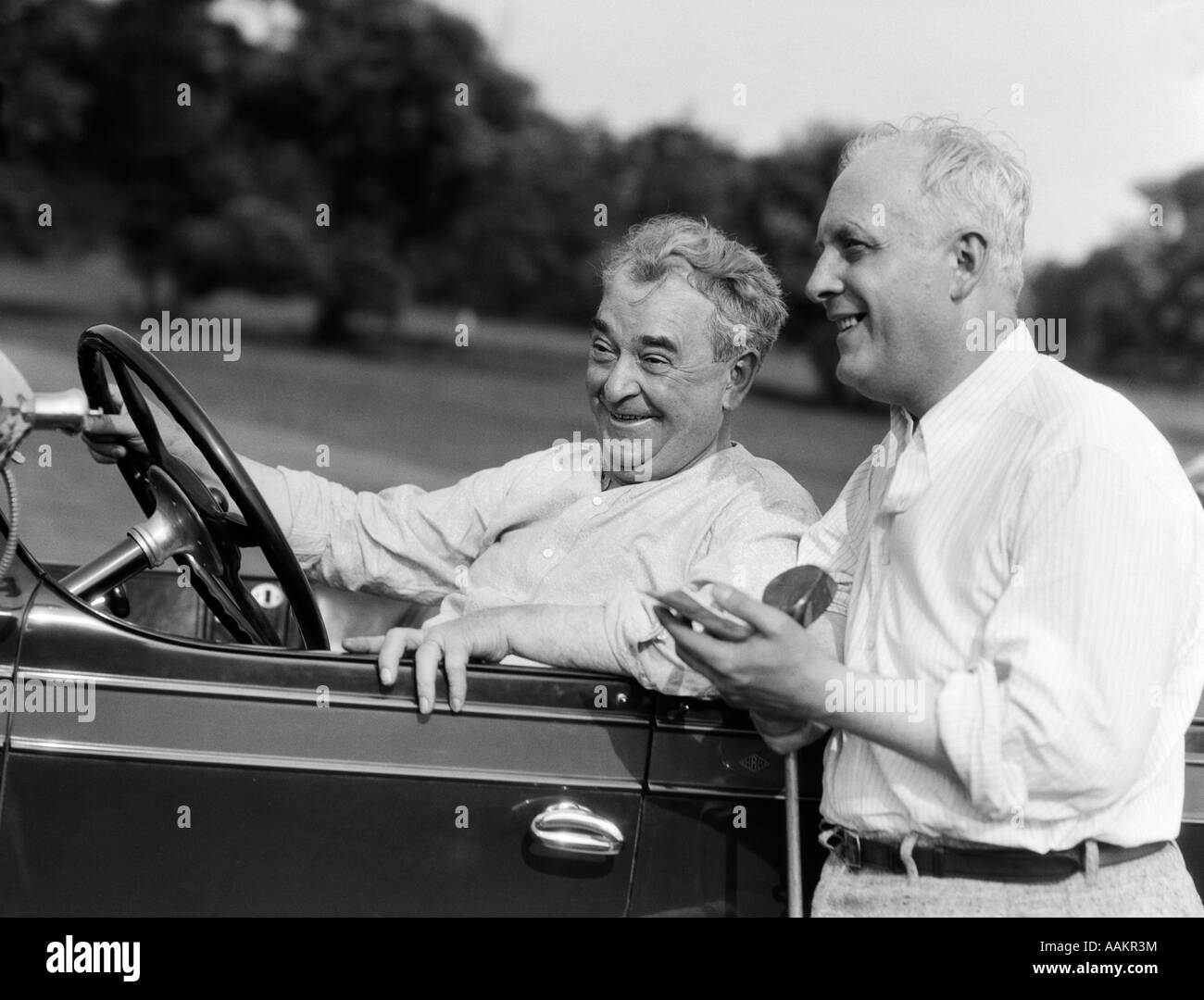 1920s 1930s SENIOR MAN SITTING DRIVING CAR AND ANOTHER STANDING HOLDING GOLF CLUB SHARING A LAUGH Stock Photo