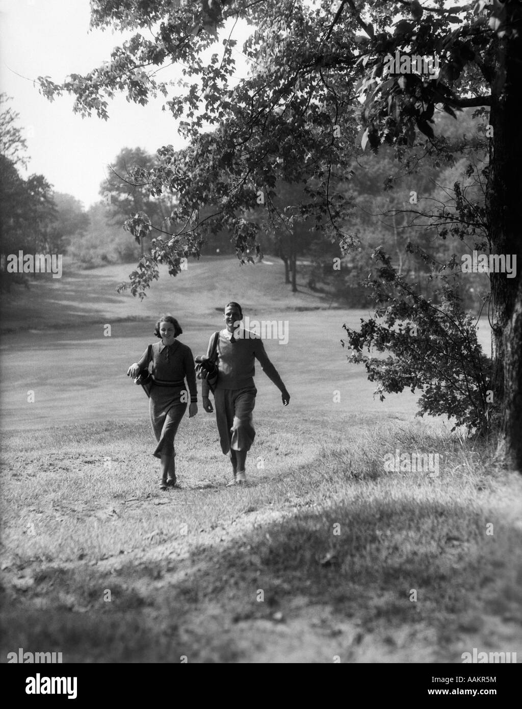 1930s COUPLE MAN WOMAN EACH CARRYING A GOLF BAG WALKING DOWN THE FAIRWAY OF THE GOLF COURSE Stock Photo