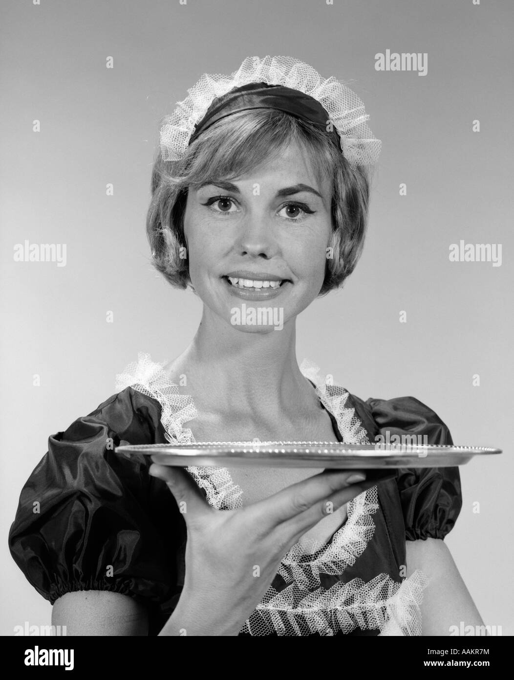 1960s GIRL IN WAITRESS UNIFORM WITH MATCHING LACE HEADPIECE SMILING WHILE HOLDING UP AN EMPTY SERVING TRAY Stock Photo