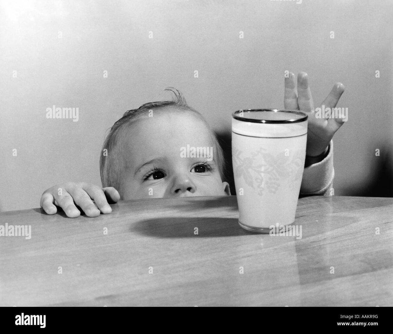 1950s TODDLER REACHING UP TO GRAB GLASS OF MILK ON TOP OF TABLE Stock Photo