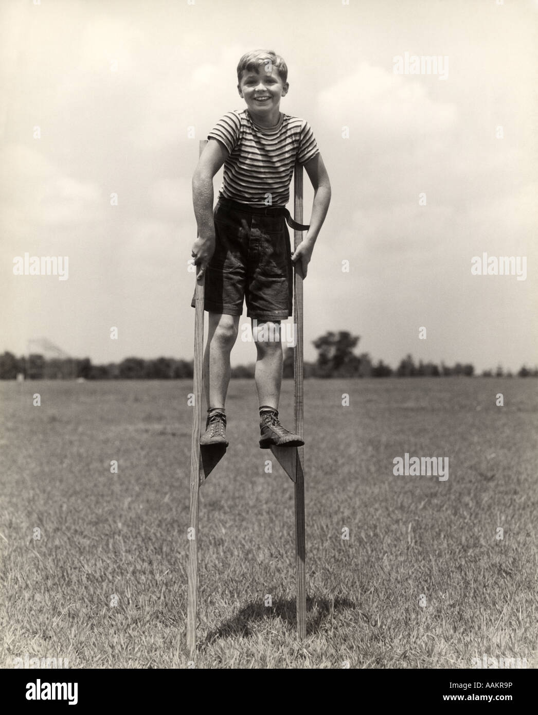 1930s 1940s SMILING HAPPY BOY WEARING STRIPED SHIRT & SHORT PANTS WALKING ON PAIR OF STILTS LOOKING AT CAMERA Stock Photo