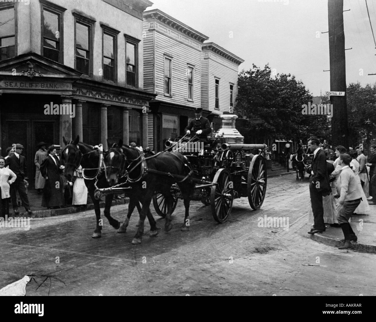 TURN OF THE CENTURY HORSE-DRAWN FIRE TRUCK PARADING THROUGH TOWN WITH PEOPLE LINING STREETS Stock Photo