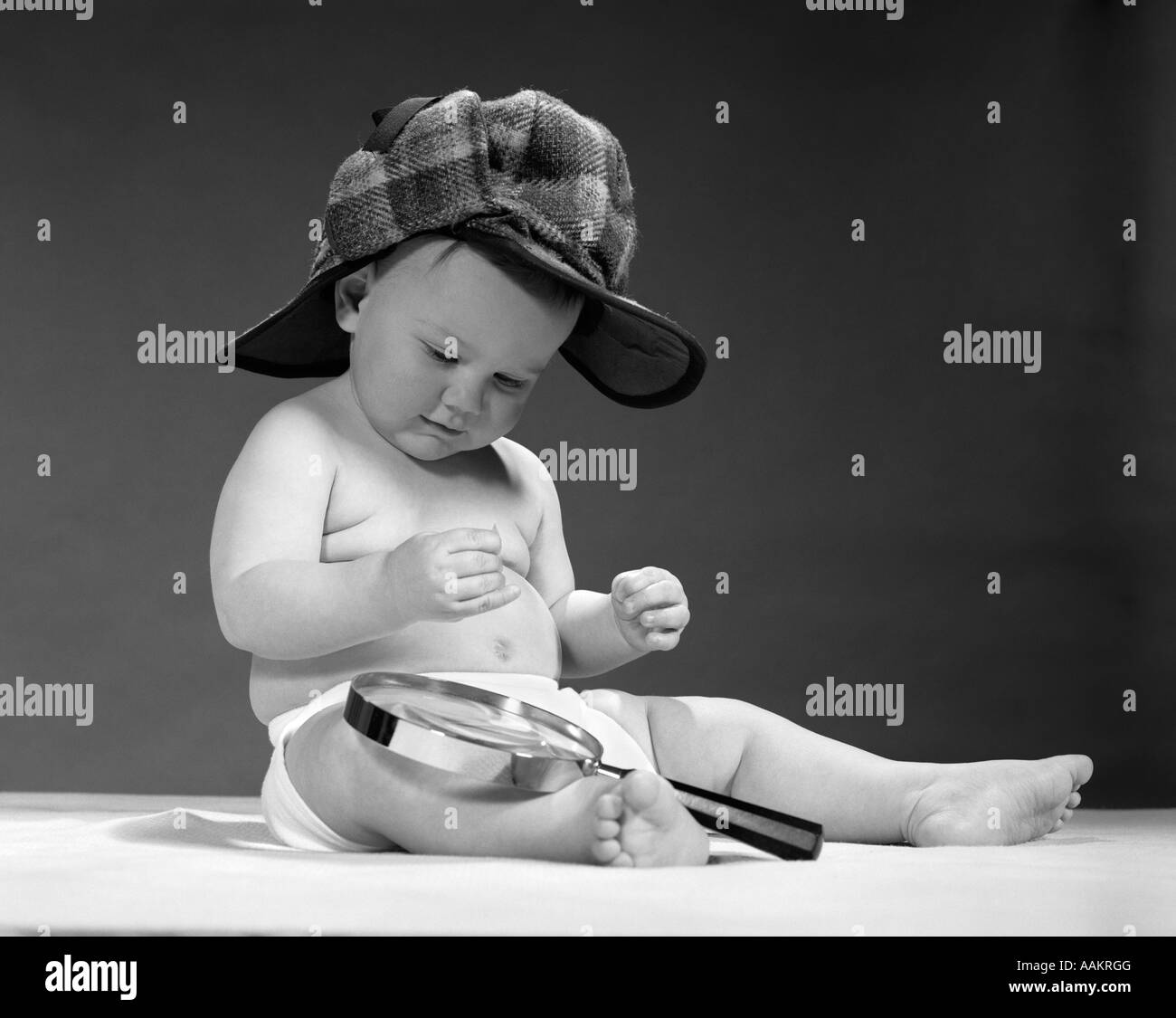 1960s BABY WEARING SHERLOCK HOLMES DEERSTALKER STYLE DOUBLE-BILLED CAP WITH MAGNIFYING GLASS RESTING ON LEG INSPECTING A CLUE Stock Photo