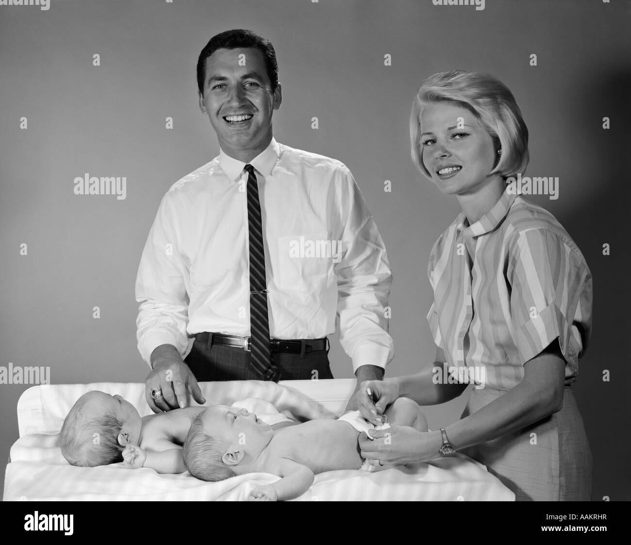 1960s SMILING MOTHER AND FATHER WITH TWIN BABIES ON DIAPER CHANGING TABLE LOOKING AT CAMERA Stock Photo