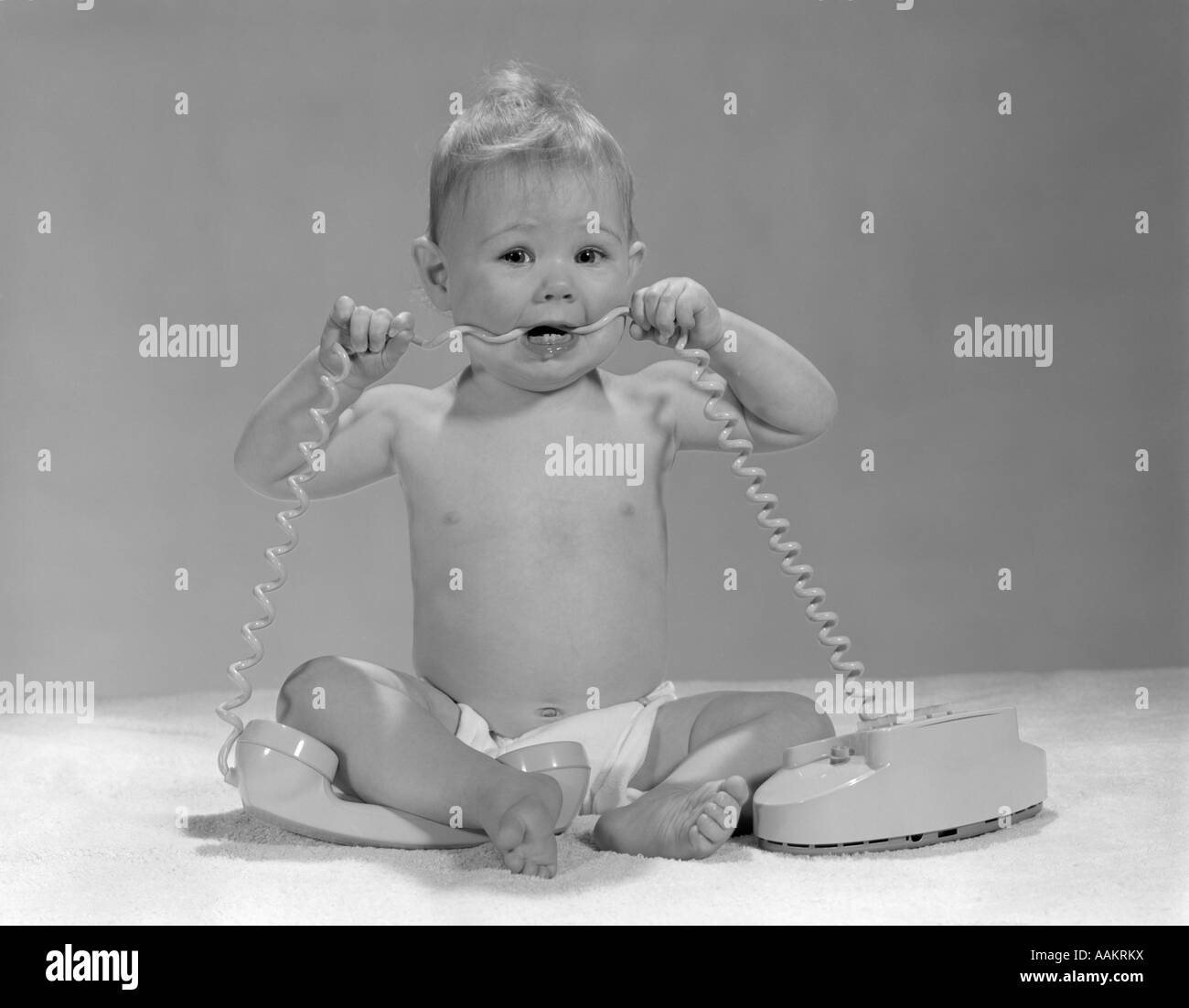 1960s BLOND BABY SITTING UP LOOKING AT CAMERA CHEWING ON TELEPHONE CORD Stock Photo