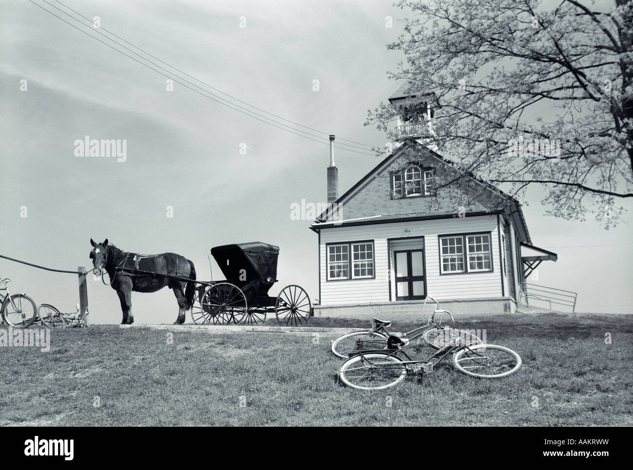 1970s AMISH ONE-ROOM SCHOOLHOUSE AT TOP OF HILL HORSE & BUGGY & BICYCLES PARKED OUTSIDE Stock Photo