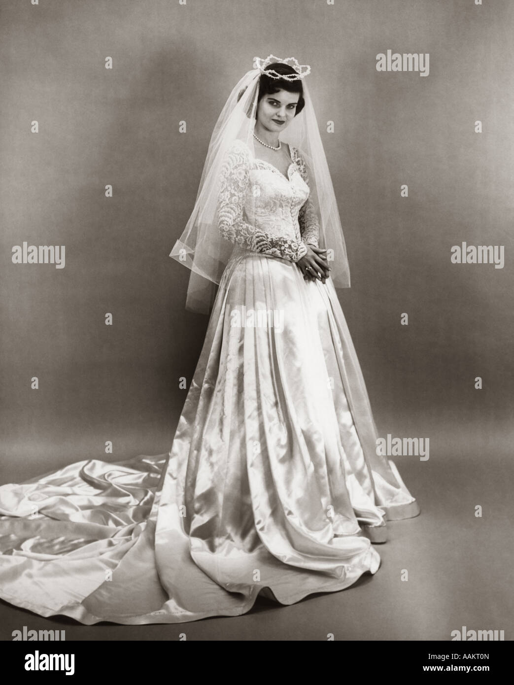 1950s FULL LENGTH PORTRAIT BRIDE STANDING WEARING SATIN AND LACE WEDDING GOWN VEIL AND TIARA LOOKING AT CAMERA Stock Photo