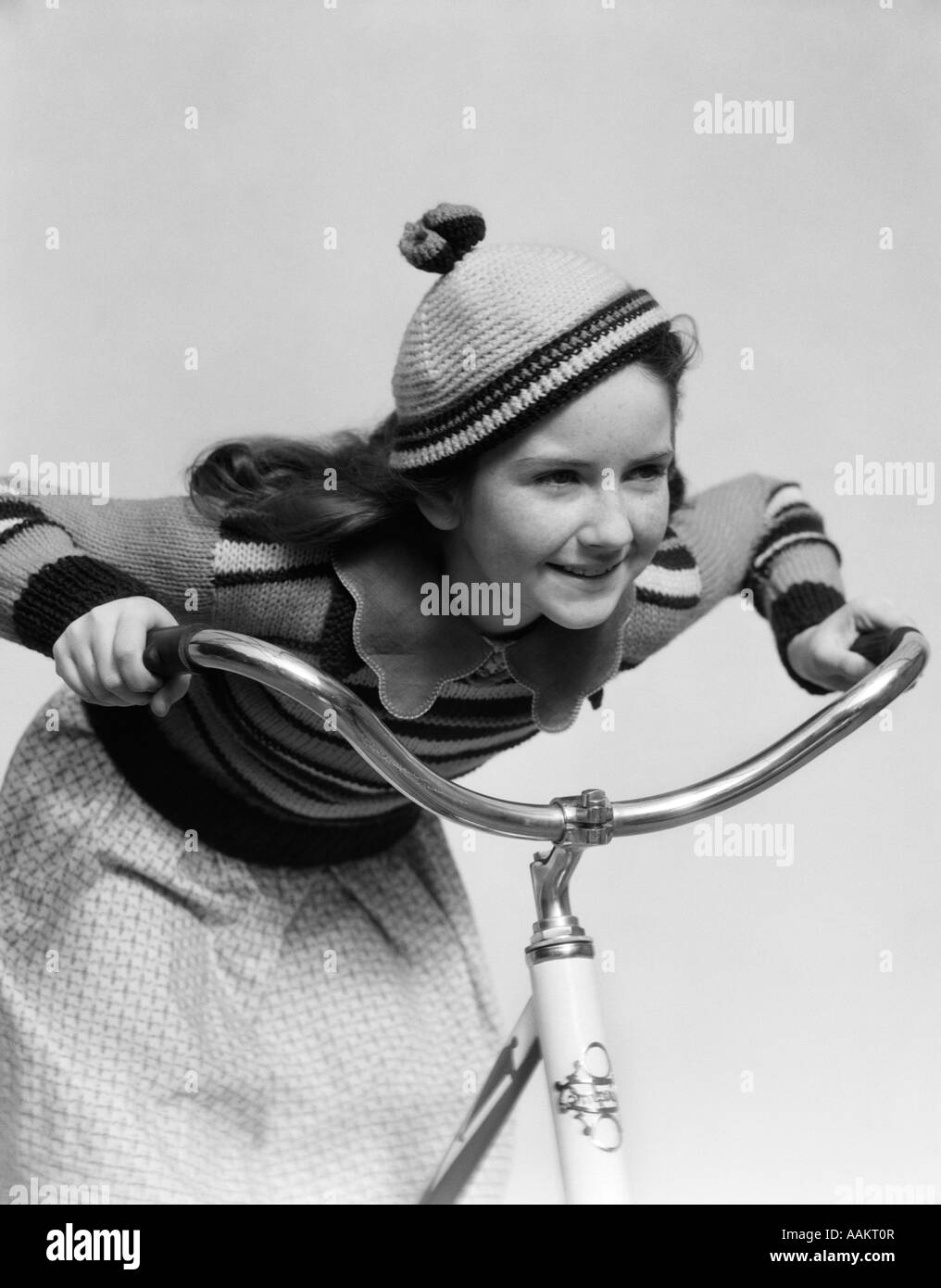 1930s SMILING EAGER LITTLE GIRL IN KNIT CAP AND MATCHING SWEATER RIDING BIKE LEANING INTO HANDLEBARS Stock Photo