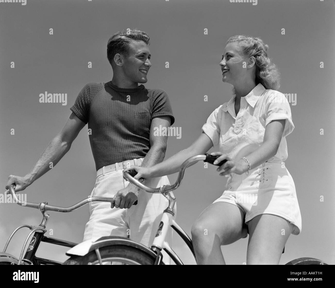 1930s 1940s SMILING BLONDE COUPLE ON BIKES LOOKING AT ONE ANOTHER Stock Photo