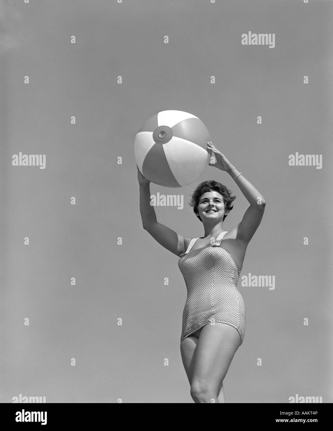 1950s SMILING WOMAN IN ONE PIECE BATHING SUIT HOLDING BEACH BALL ABOVE HEAD LOOKING Stock Photo