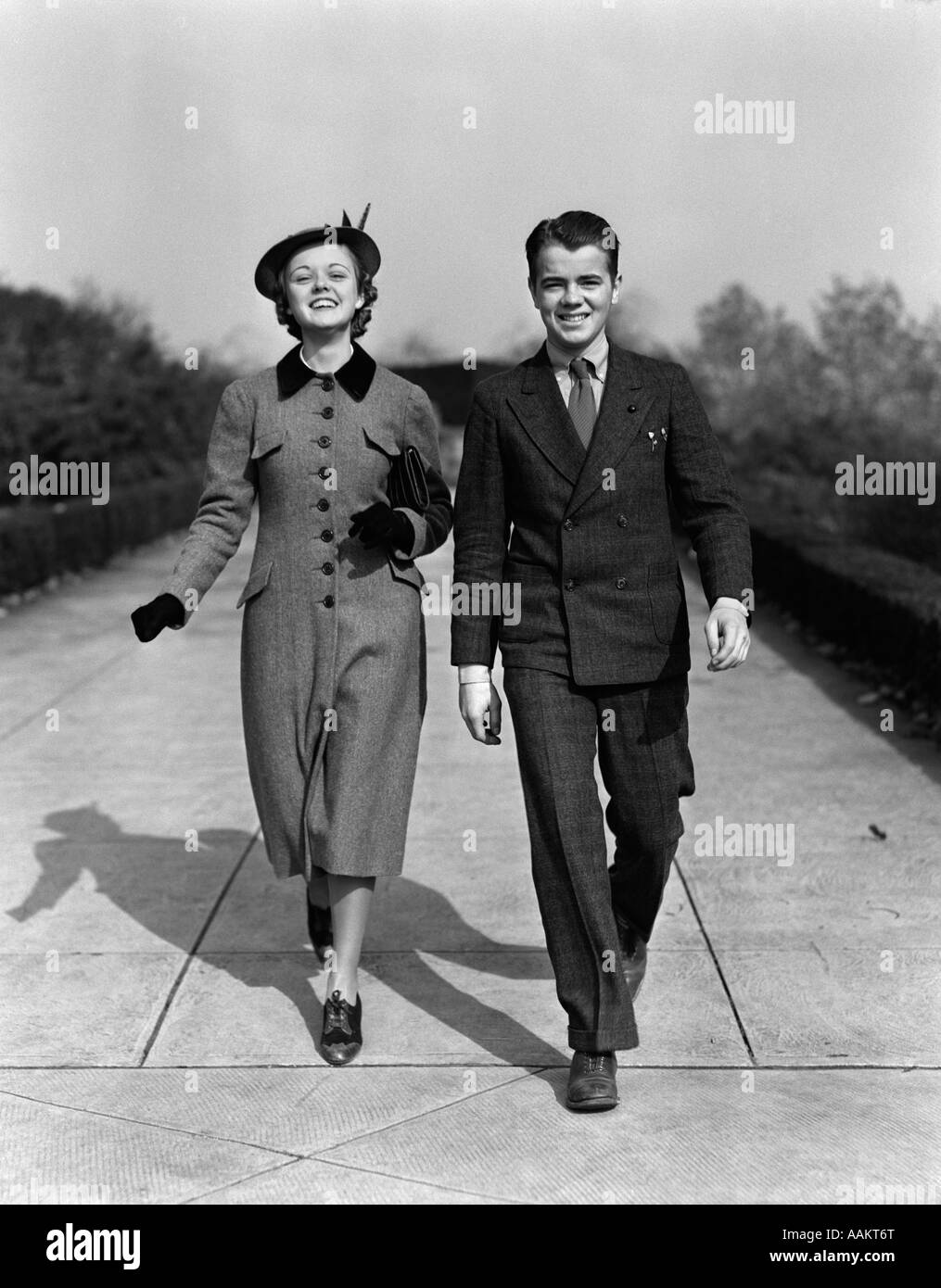 1940s YOUNG SMILING TEENAGE COUPLE WALKING ON SIDEWALK SMILING DRESSED UP SUNDAY BEST Stock Photo