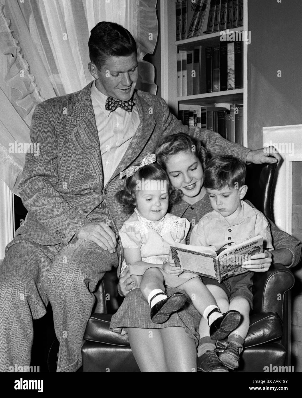 1940s WOMAN MOTHER IN ARMCHAIR READING TO BOY SON GIRL DAUGHTER WITH PROUD MAN FATHER WATCHING Stock Photo