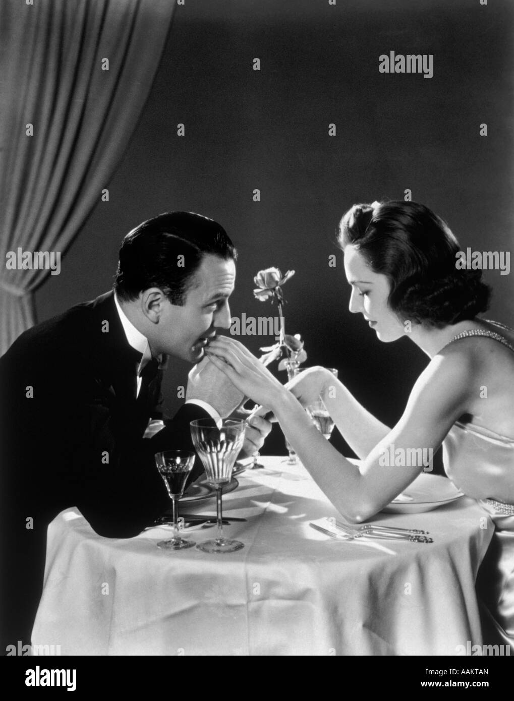 1930s ROMANTIC COUPLE AT DINING TABLE MAN KISSING WOMAN'S HAND Stock Photo