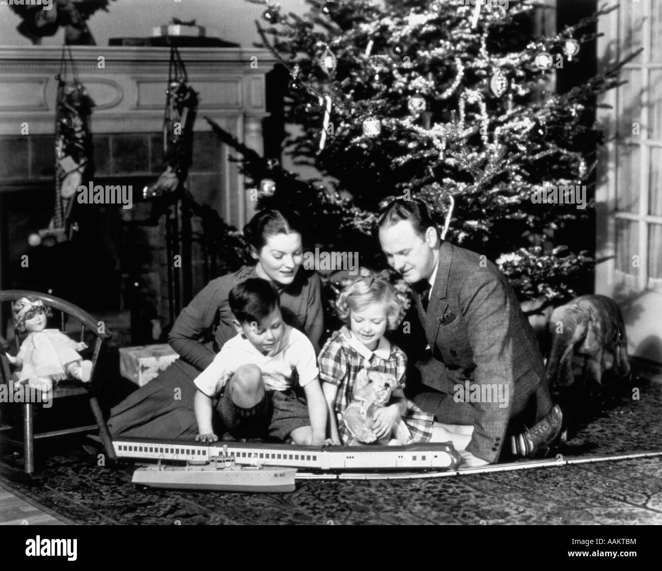 1930s FAMILY PLAYING WITH TOY TRAIN IN FRONT OF CHRISTMAS TREE FIREPLACE Stock Photo
