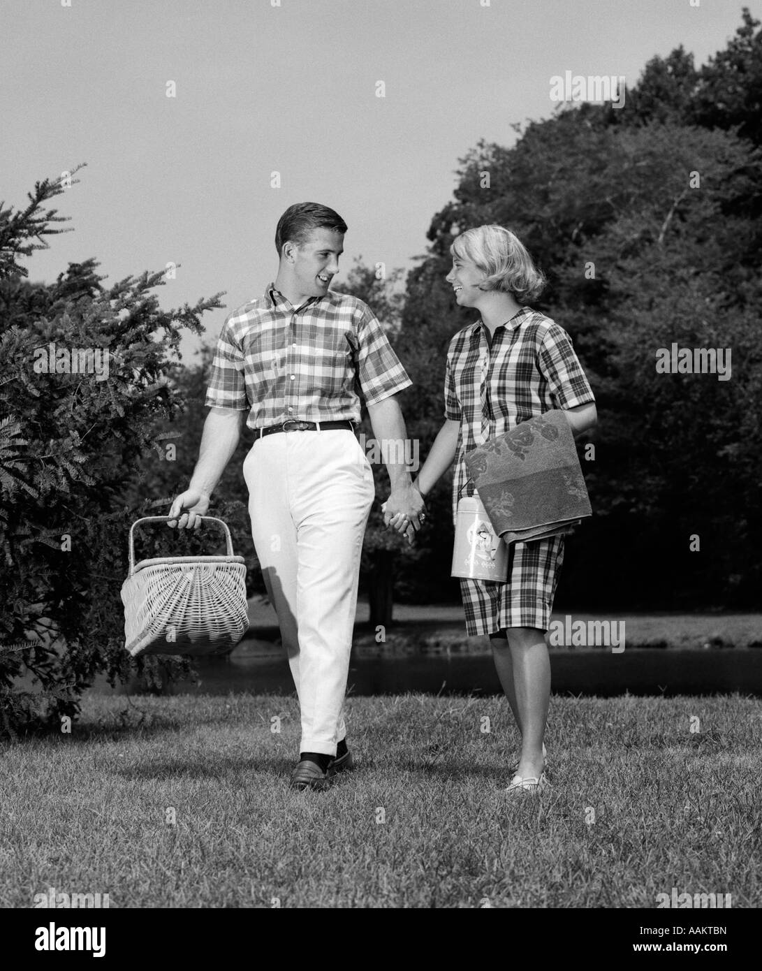 1960s TEENAGE COUPLE HOLDING HANDS WALKING WITH PICNIC BASKET TALKING FLIRTING DATING Stock Photo