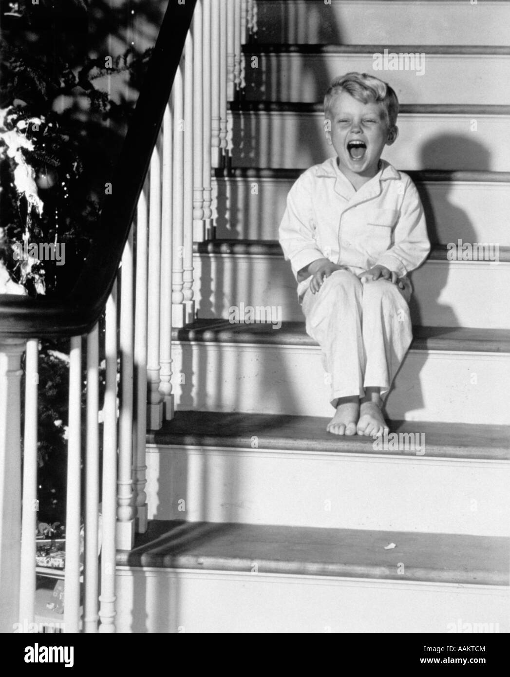 1950s LAUGHING HAPPY LITTLE BOY IN PAJAMAS SITTING ON STAIRS CHRISTMAS MORNING Stock Photo