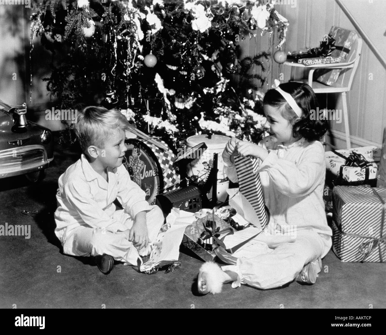1950s BOY AND GIRL SITTING UNDER CHRISTMAS TREE OPENING PRESENTS Stock Photo