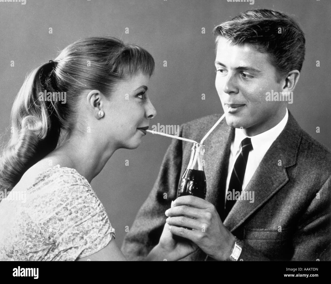 1950s TEENAGE COUPLE SHARING A SINGLE BOTTLE OF SODA WITH TWO STRAWS Stock Photo