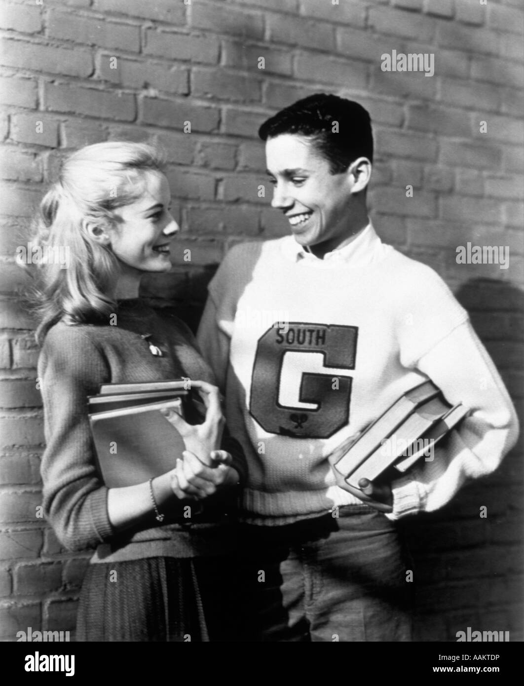 1950s TEENAGE COUPLE HOLDING BOOKS AND SMILING LEANING AGAINST WALL BOY WEARS VARSITY LETTER SWEATER Stock Photo