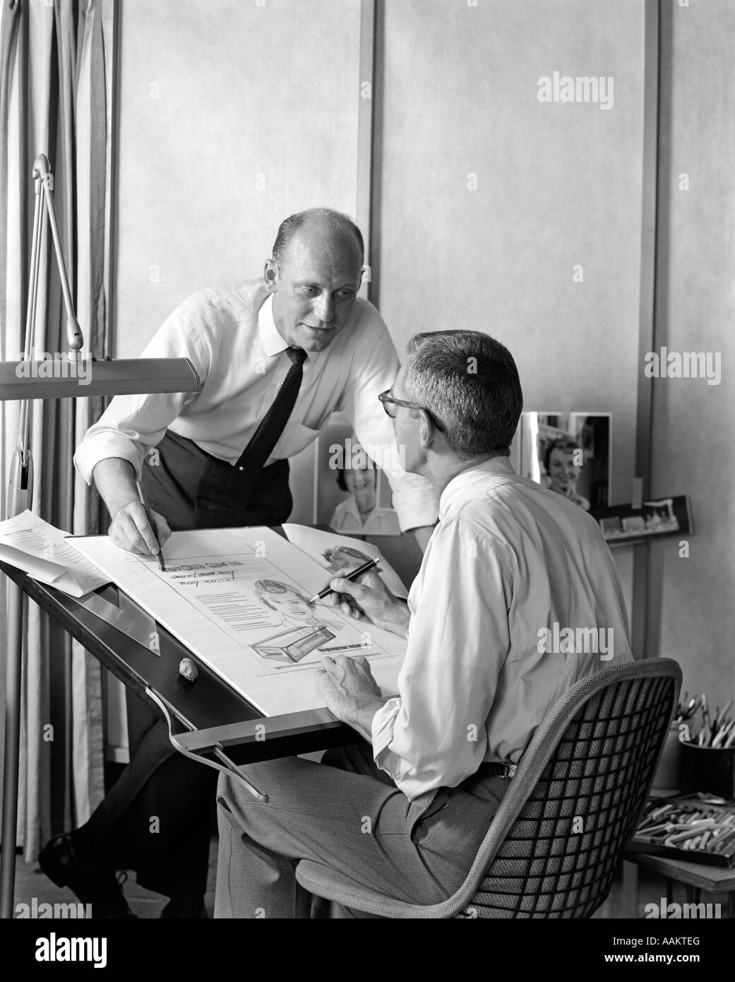 1950s 1960s TWO ADVERTISING AGENCY MEN AT ARTIST DRAFTING TABLE REVIEWING AN AD Stock Photo