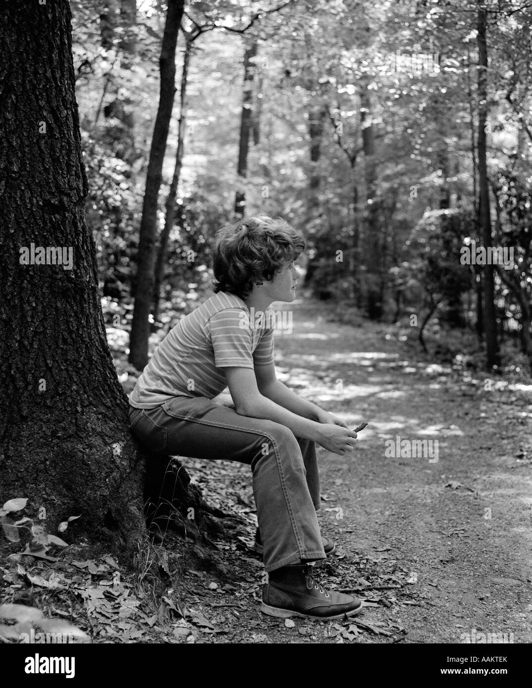 1970s SIDE VIEW OF BOY SITTING AT FOOT OF TREE ALONG TRAIL IN WOODS Stock Photo