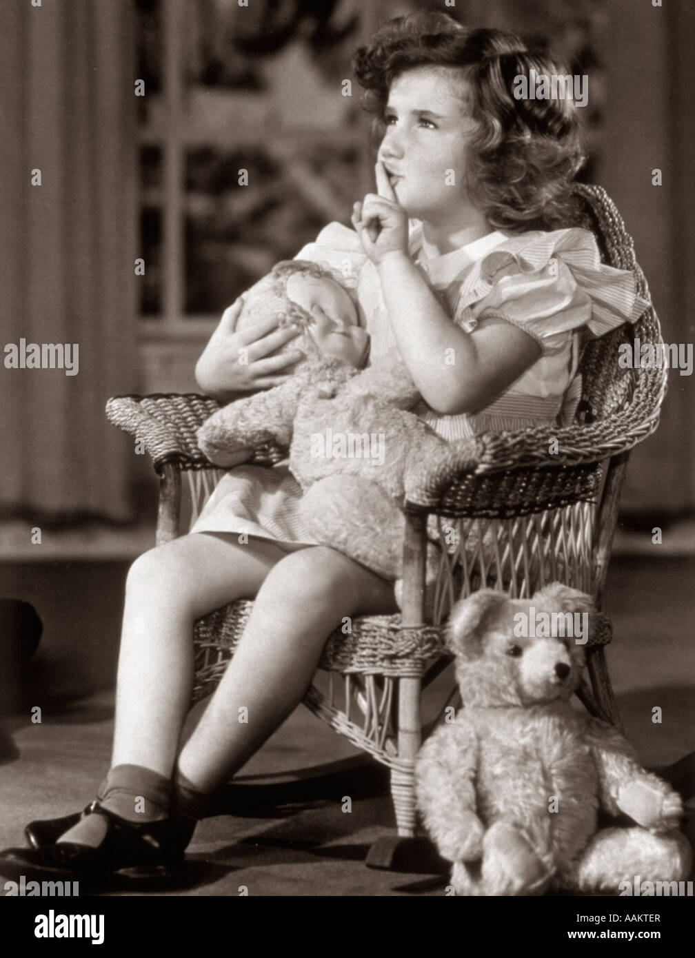 1930s 1940s GIRL MAKING SHUSHING SIGN AS SHE ROCKS HER DOLL TO SLEEP IN WICKER ROCKING CHAIR Stock Photo