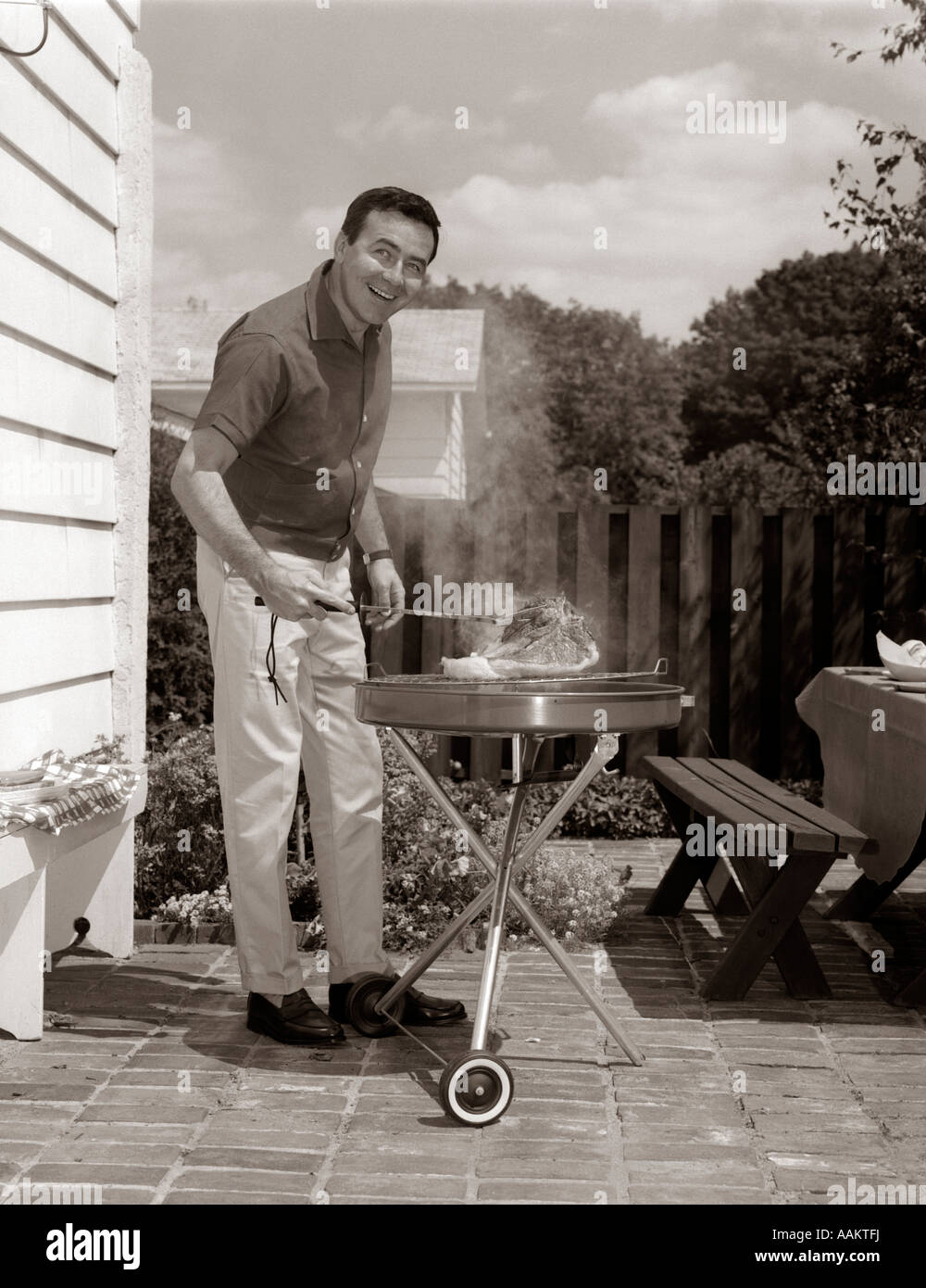1960s HEAD-ON VIEW OF MAN BACKYARD PATIO COOKING STEAK ON GRILL Stock Photo