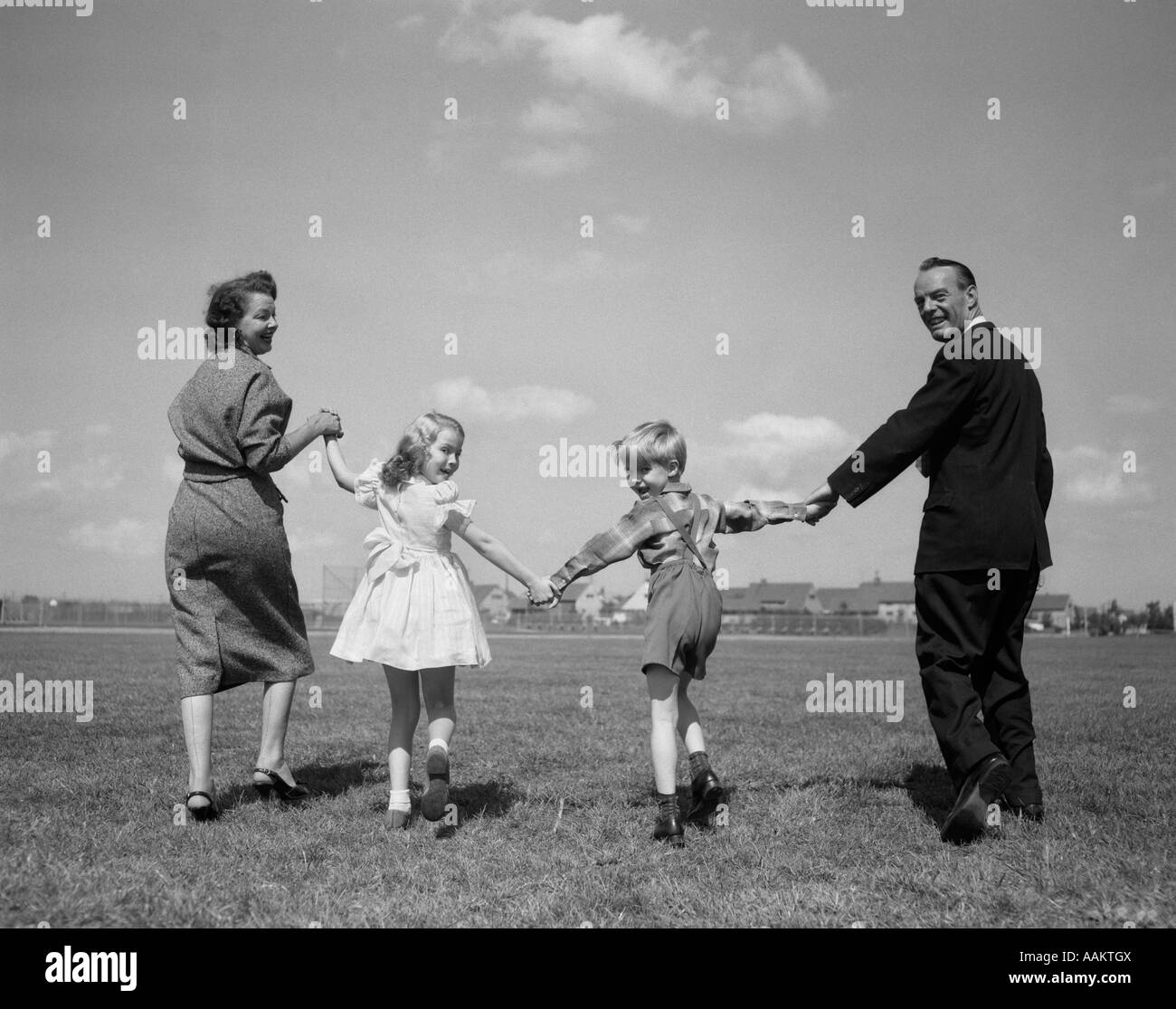 1950s SMILING HAPPY FAMILY MOTHER FATHER BOY GIRL HOLDING HANDS WALKING AWAY ON GRASS TURNED BACK LOOKING AT CAMERA Stock Photo