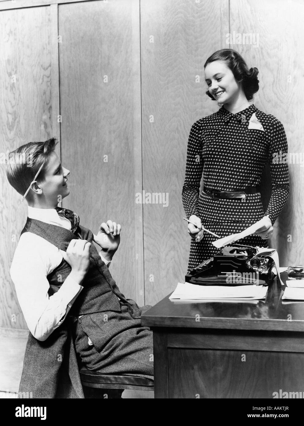 1940s TEEN YOUNG COUPLE BOY AT DESK AS BUSINESSMAN GIRL POLKA DOT DRESS STANDING AS SECRETARY SCHOOL PAPER Stock Photo