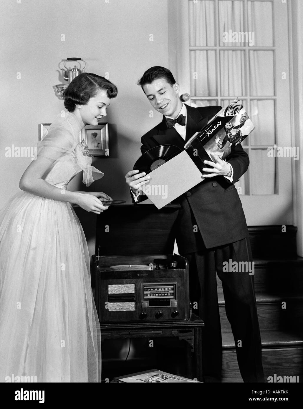 1950s TEEN COUPLE BOY AND GIRL IN PROM FORMAL WEAR PLAYING PHONOGRAPH RECORDS IN HOME LIVING ROOM Stock Photo
