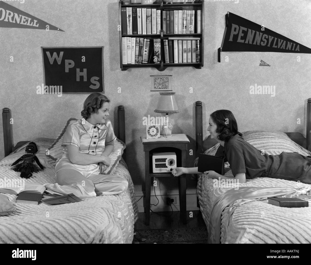 1930s 1940s TWO TEEN GIRLS LYING ON DORMITORY BEDS ROOM MATES LISTENING TO RADIO COLLEGE SCHOOL PENNANTS ON WALL Stock Photo