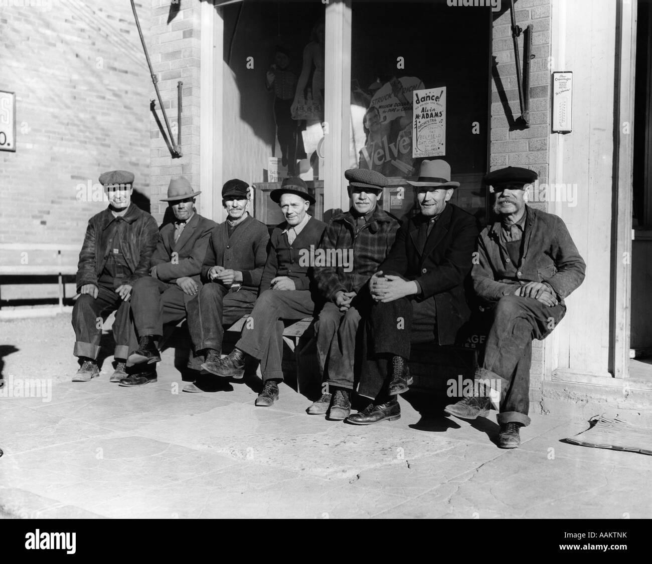 1920s 1930s GROUP OF OLD MEN IN HATS & DUNGAREES SITTING ON BENCH IN FRONT OF STORE WINDOW LOOKING AT CAMERA Stock Photo