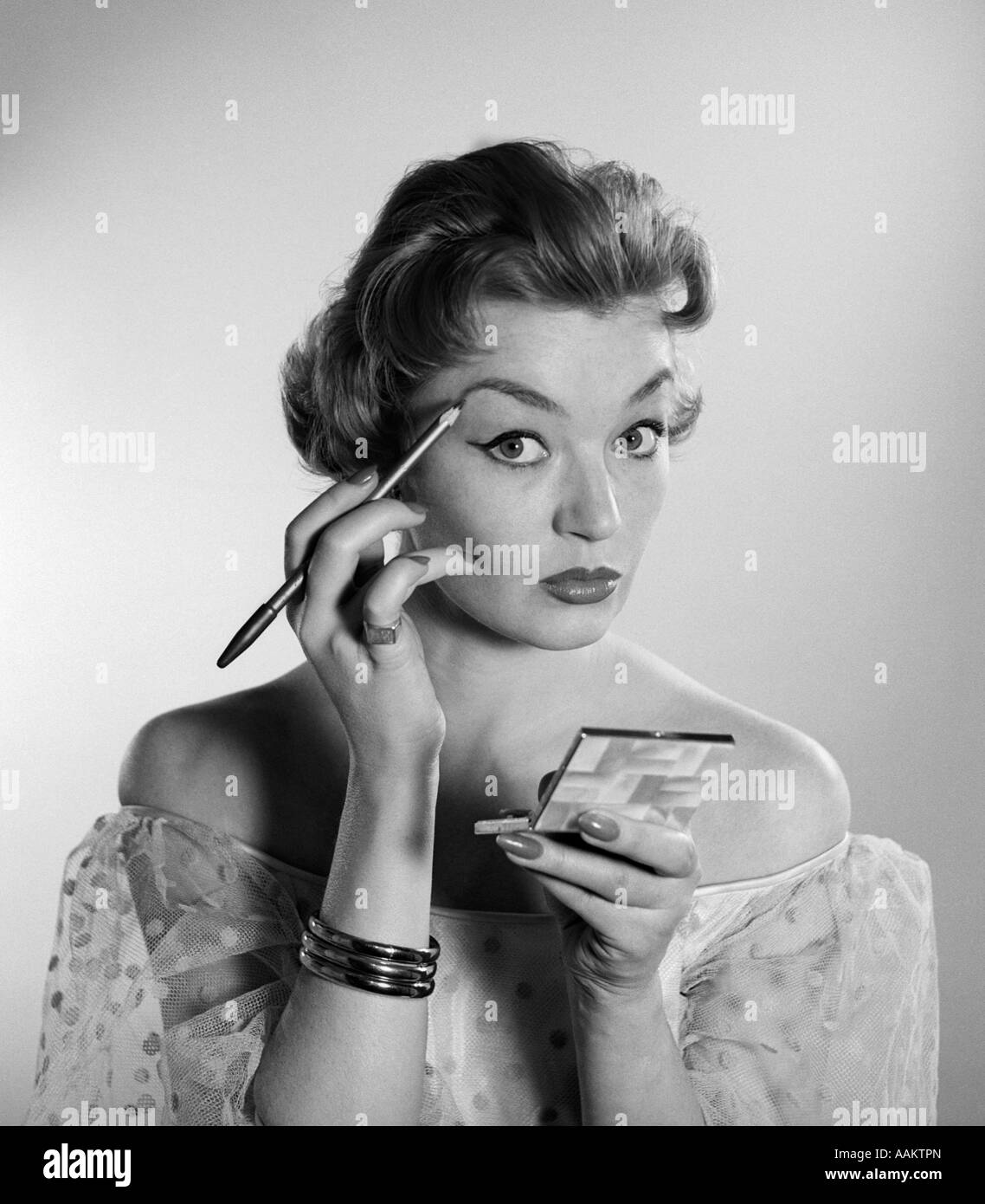 1950s 1960s WOMAN LOOKING AT CAMERA APPLYING MAKEUP EYE BROW PENCIL HOLDING COMPACT MIRROR Stock Photo