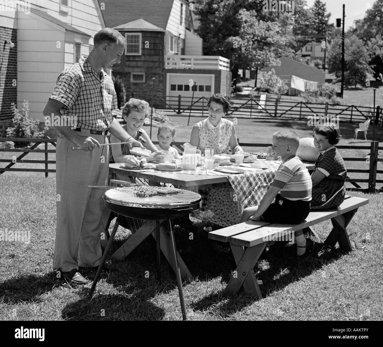 1950s FAMILY IN BACKYARD COOKING HOT DOGS SITTING AT PICNIC TABLE Stock Photo