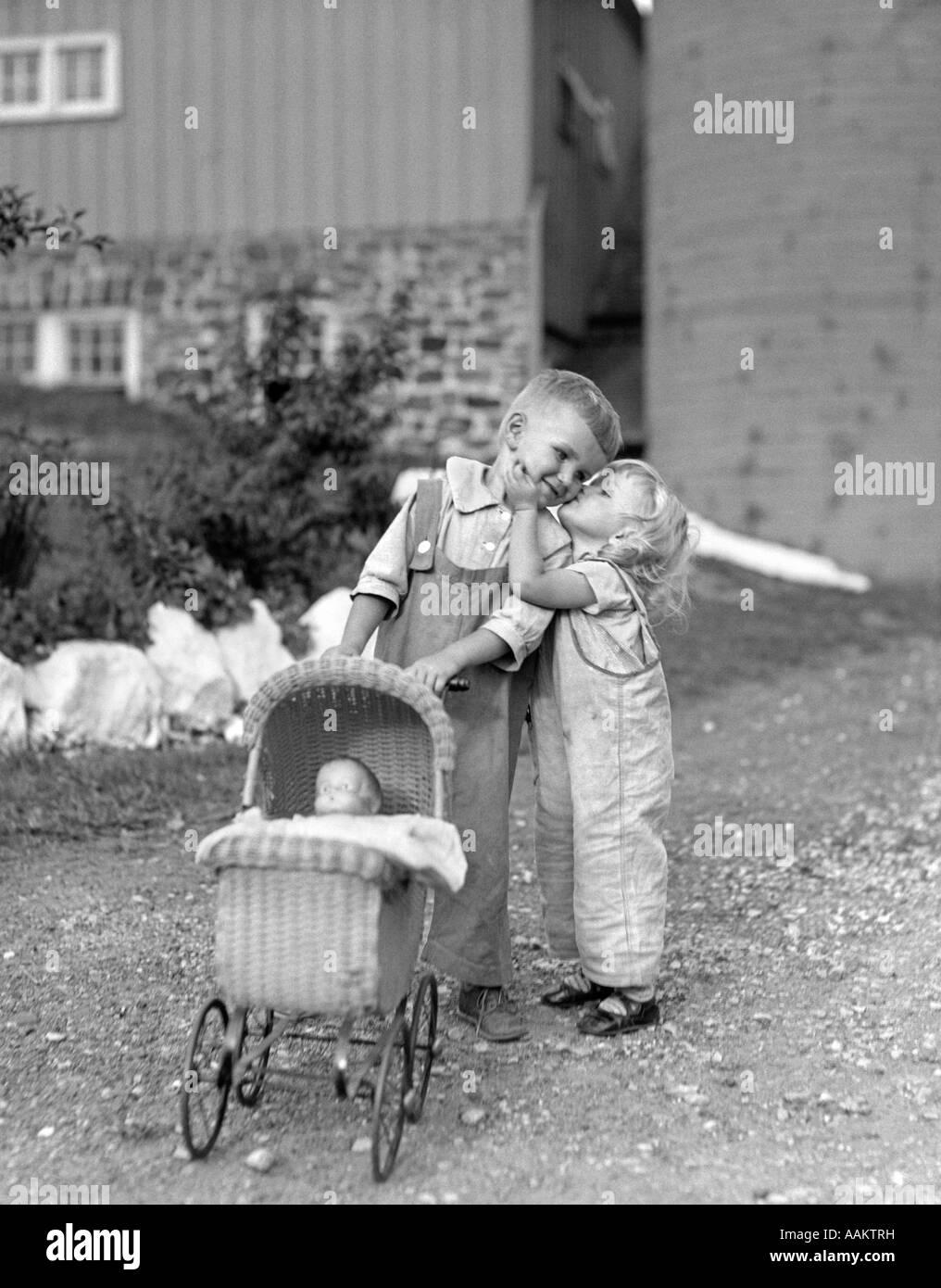 1930s 1940s LITTLE GIRL KISSING BOY PUSHING TOY BABY CARRIAGE IN FRONT OF BARN Stock Photo