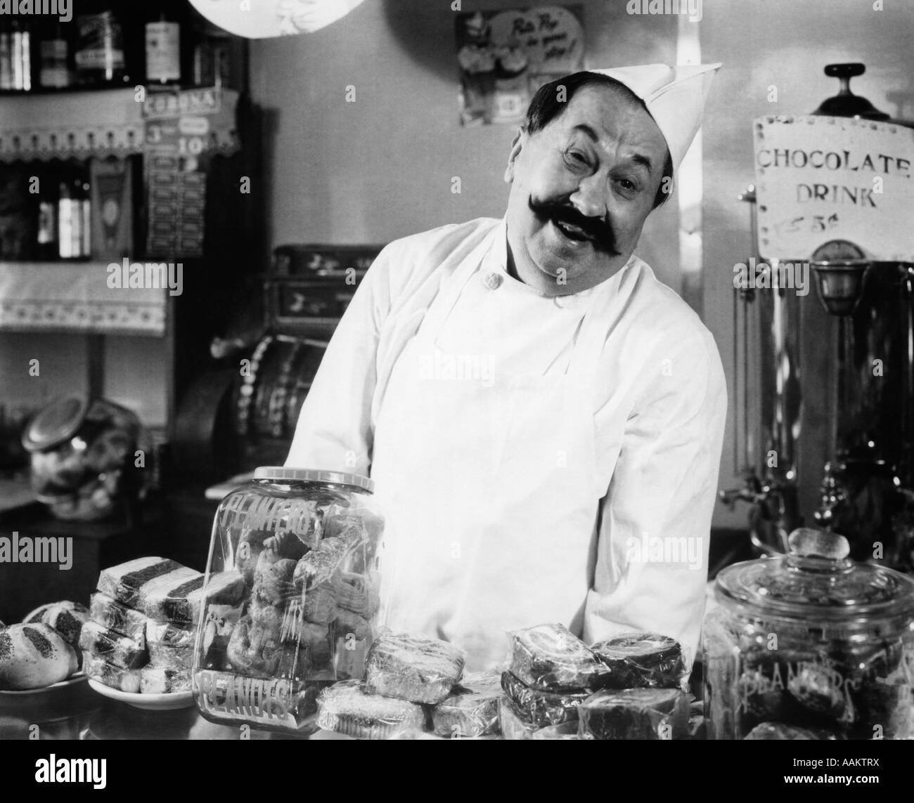 1930s VINTAGE PORTRAIT OF SMILING PROPRIETOR BEHIND COUNTER IN COFFEE AND SWEET SHOP LOOKING AT CAMERA Stock Photo