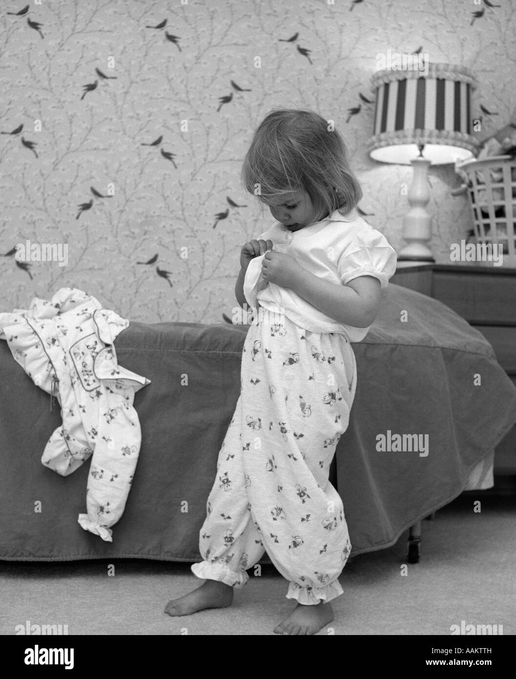 1960s BEDTIME LITTLE BLOND GIRL IN PAJAMA BOTTOMS UNBUTTONING SHIRT TO PUT ON PAJAMA TOP Stock Photo
