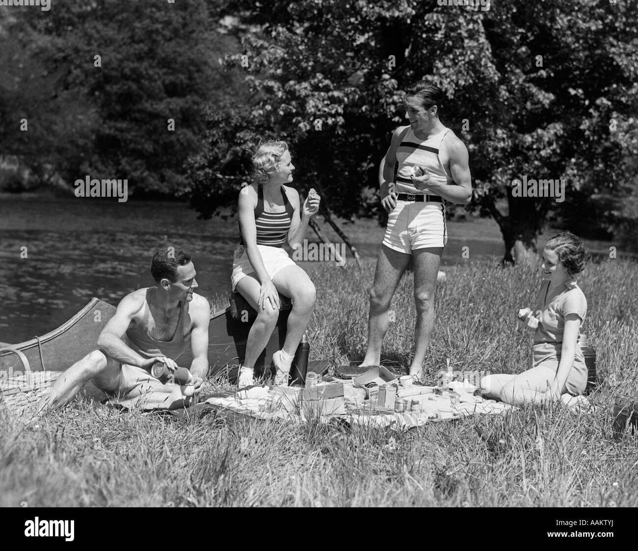 1930s TWO COUPLES HAVING SUMMER PICNIC WITH FOOD AND DRINK SPREAD OUT ON BLANKET THE TIP OF A CANOE IS VISIBLE Stock Photo