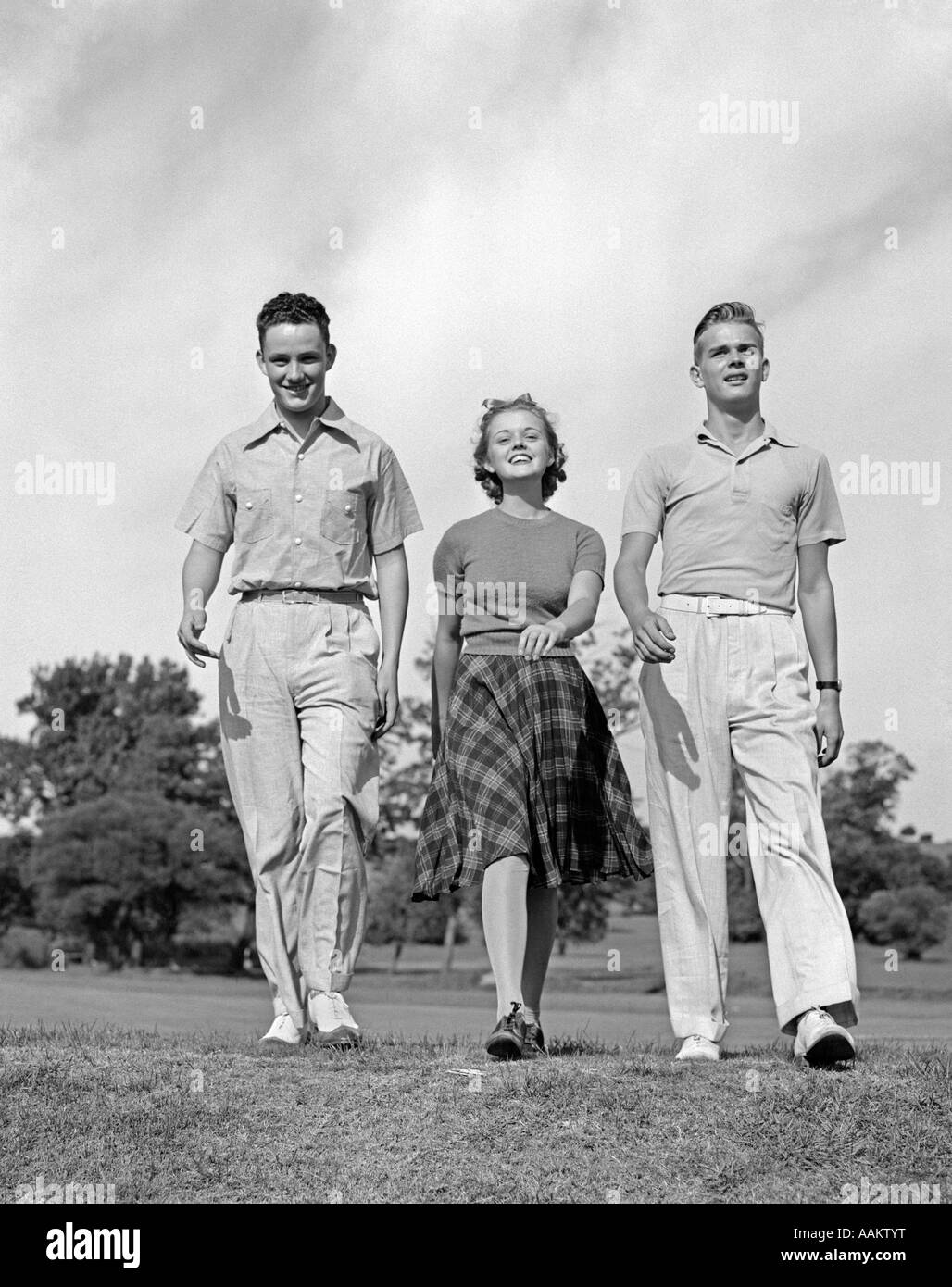 1930s 1940s TWO TEEN BOYS ONE GIRL WALKING CONFIDENTLY IN GRASSY MEADOW CASUAL DRESS Stock Photo