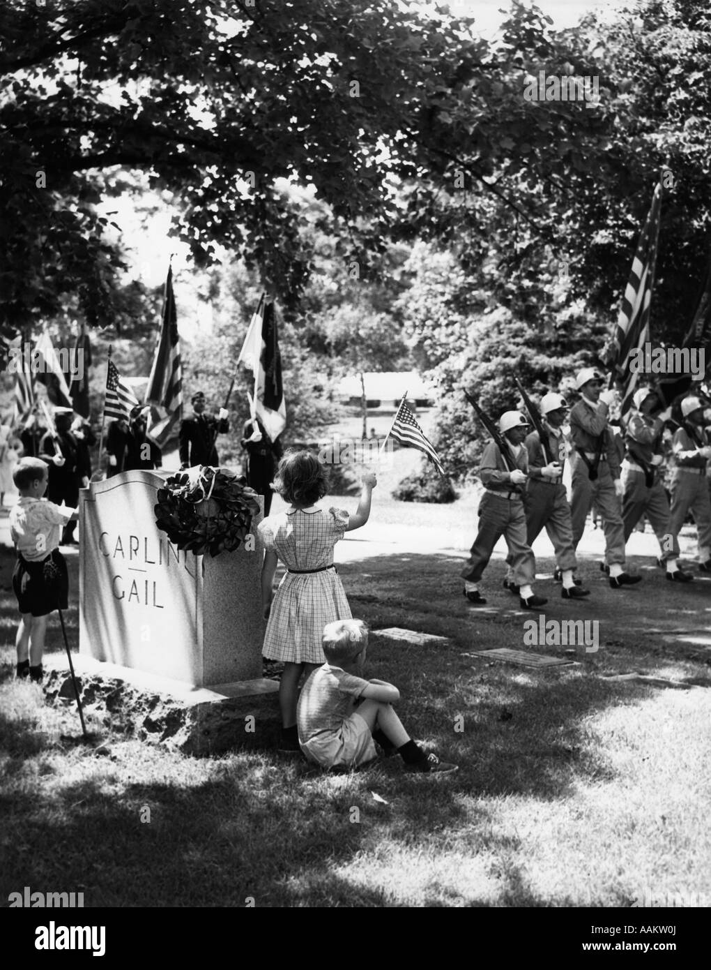 1950s A BOY AND A GIRL STAND BY A TOMBSTONE WAVING TO PASSING COLOR GUARD OF SOLDIERS IN A MEMORIAL DAY PARADE Stock Photo