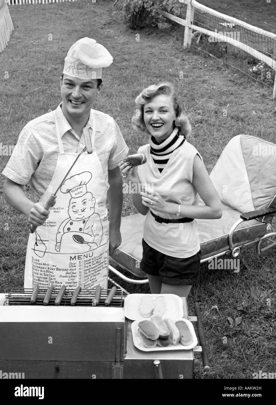 1950s COUPLE BACKYARD GRILL COOK HOT DOGS MAN WEARING APRON TOQUE & SKEWERED HOT DOG Stock Photo