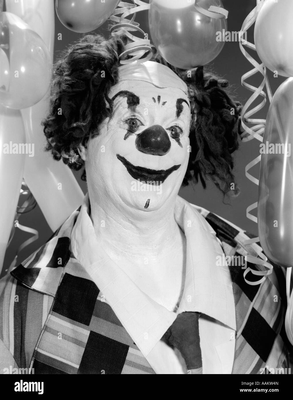1960s PORTRAIT WHITEFACE CLOWN IN FULL COSTUME SURROUNDED BY BALLOONS LOOKING AT CAMERA Stock Photo