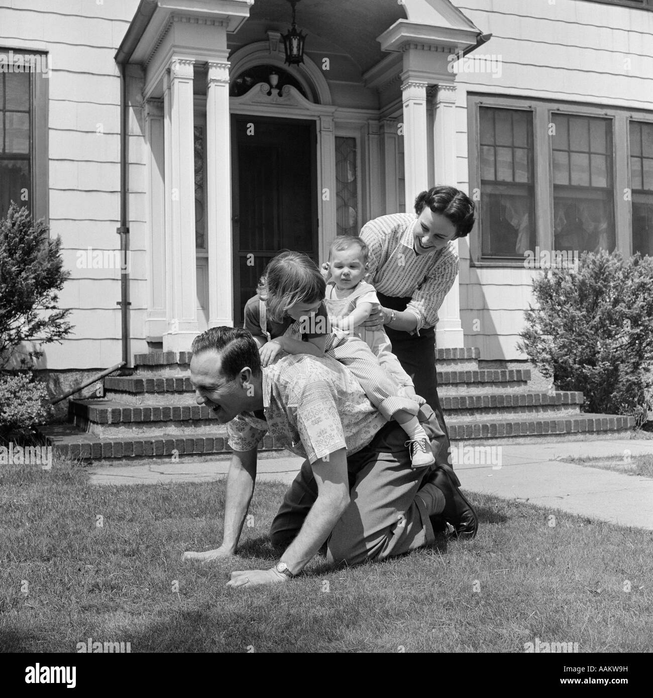 1950s FAMILY FRONT LAWN HOUSE TWO KIDS RIDING FATHER PIGGYBACK MOTHER HELPING TODDLER BABY Stock Photo