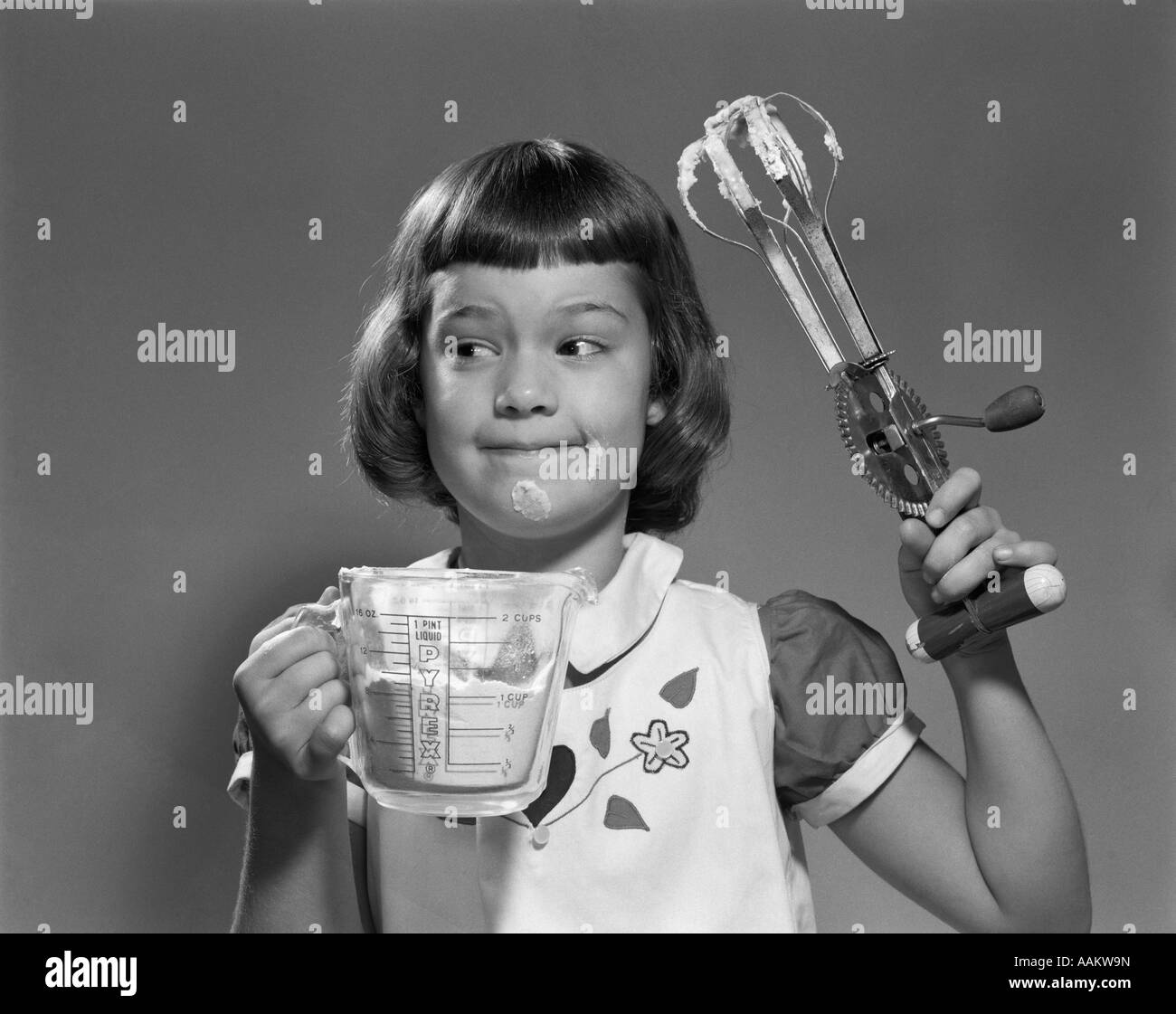 1950s SMILING GIRL HOLDING MEASURING CUP EGG BEATERS COOKING MIXING FOOD ON FACE Stock Photo
