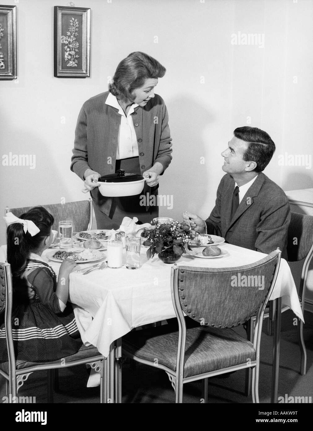 1950s 1960s FAMILY OF THREE AT DINING ROOM TABLE AT DINNER TALKING WITH WIFE STANDING PUTTING CASSEROLE DISH ON TABLE Stock Photo