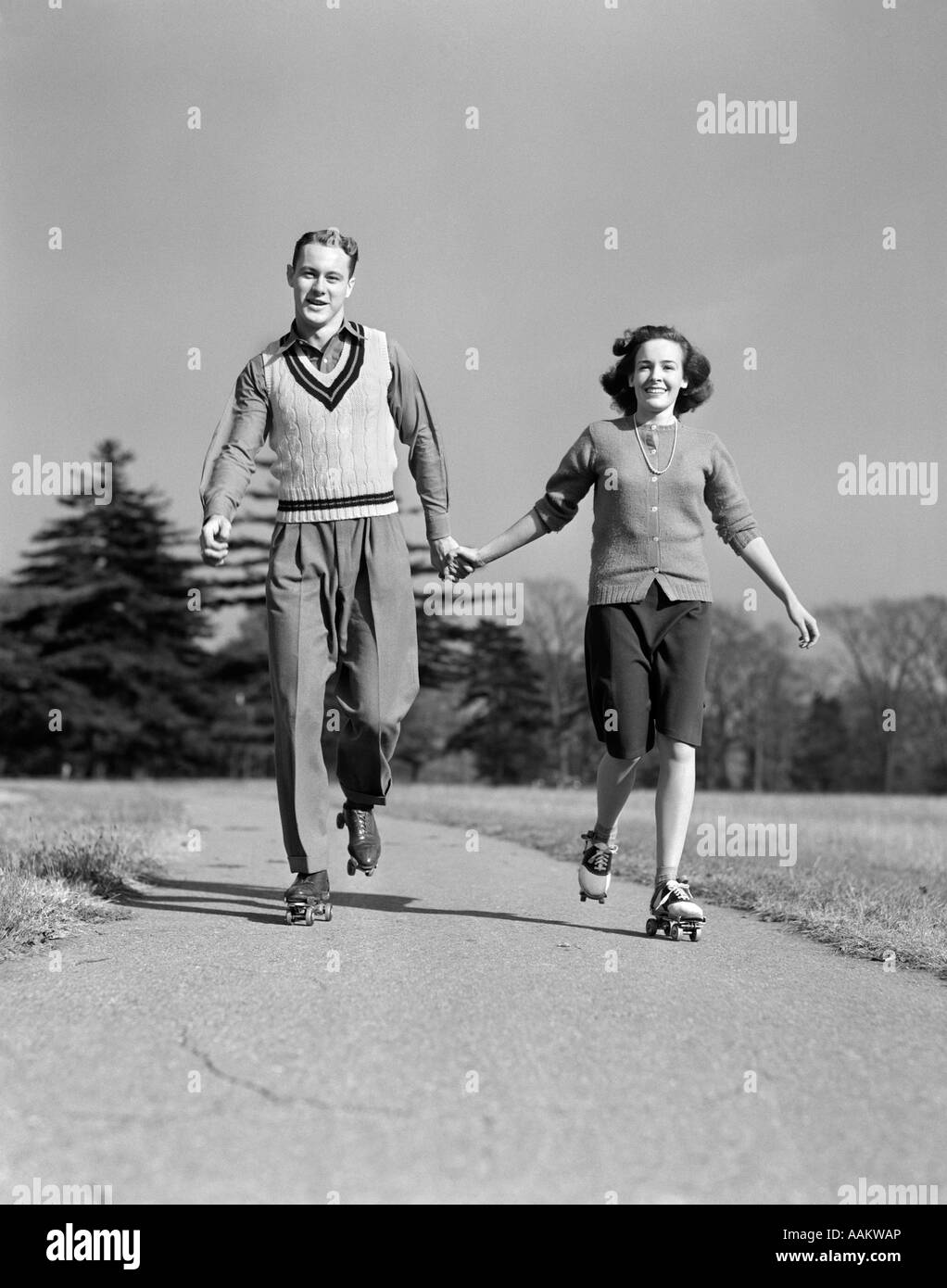 1940s SMILING TEENAGE COUPLE BOY GIRL HOLDING HANDS ROLLER SKATING ON SIDEWALK Stock Photo